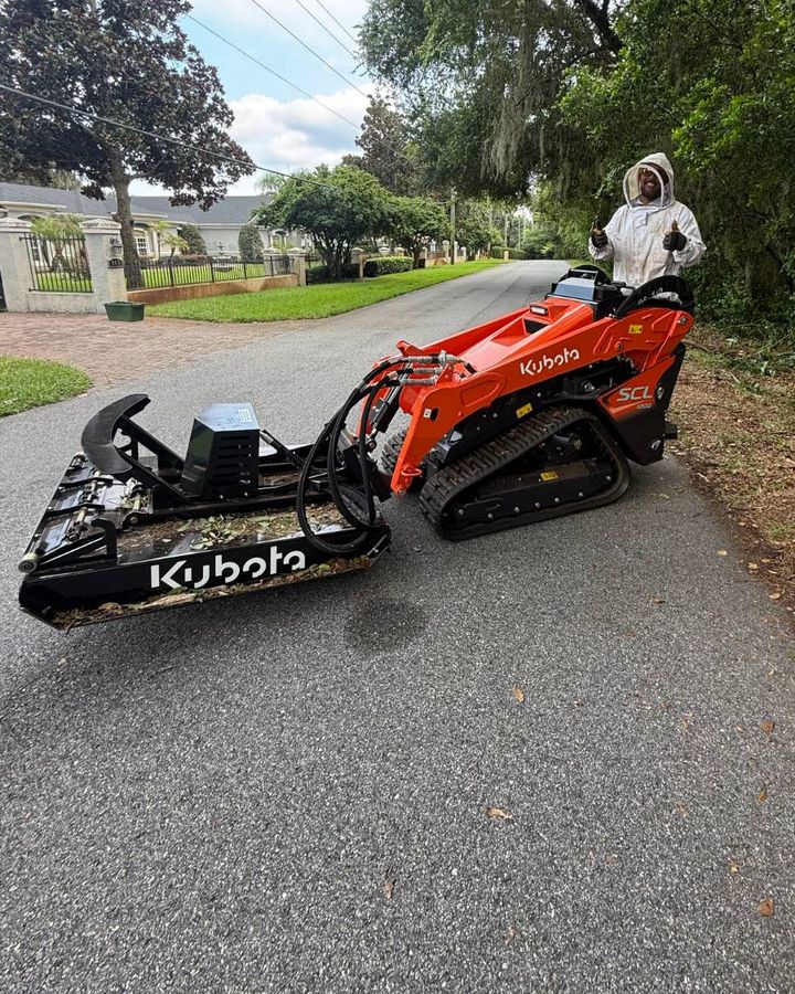 Orange Kubota SCL1000 stand-on track loader with a black brush cutter attachment on a road lined with oaks and Spanish moss. The operator, in a white protective suit, gives a thumbs up.