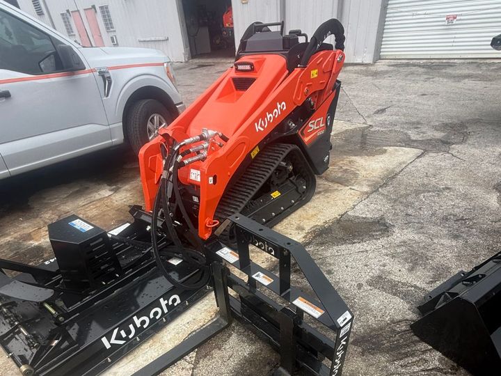 Kubota SCL1000 track loader with Kubota pallet forks on the front and the Kubota brush cutter on the ground beside it.